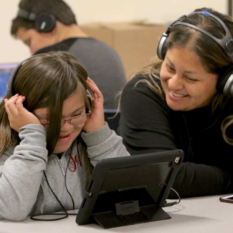 An adult and a child with headphones on sitting in a classroom looking at a tablet