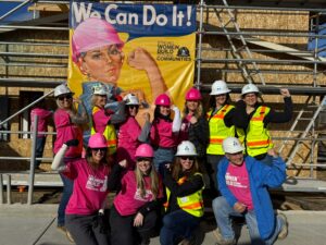Women in pink and white construction hats posing in front of a "We Can Do It!" sign for Strong Women Build Strong Communities.