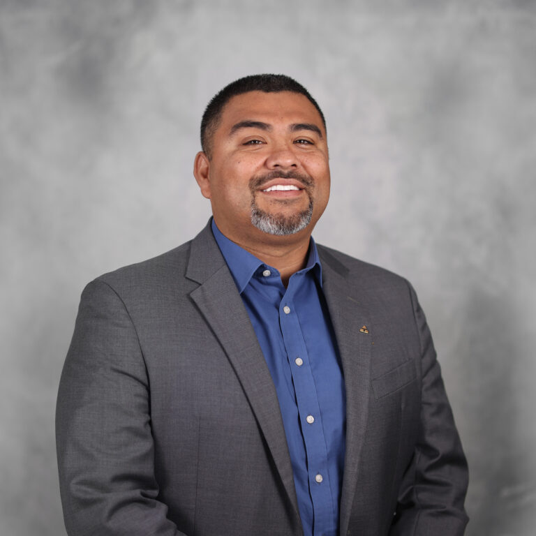 A headshot of Raymond Hernandez, a man with short dark hair wearing a dark grey shirt with a blue shirt