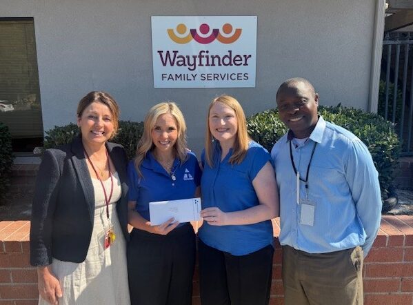 Four woman and one man posing in front of the Wayfinder Family Services sign holding an envelope.